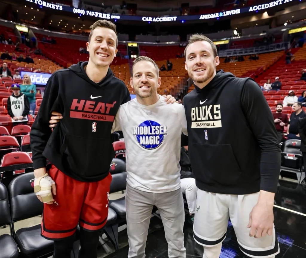 Middlesex Magic shirt courtside, flanked by two NBA players in Heat and Bucks warmups